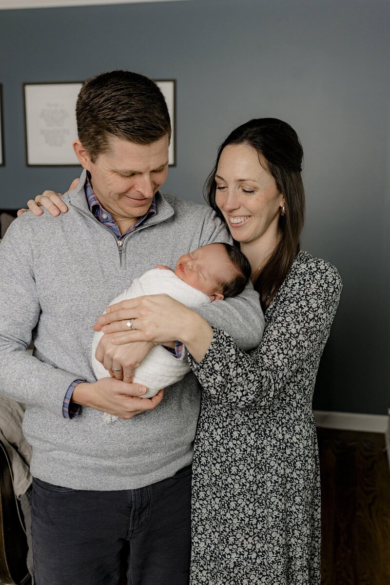 Mother and father looked down towards their newborn son as they stand and their master bedroom while their newborn is wrapped in white and laying gently and Dad's arms