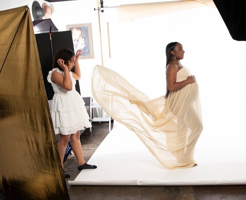 inside the studio of Insley photography pregnant mom to be in a cream fabric while her young daughter throws dress to assists by creating motion in the fabric.
