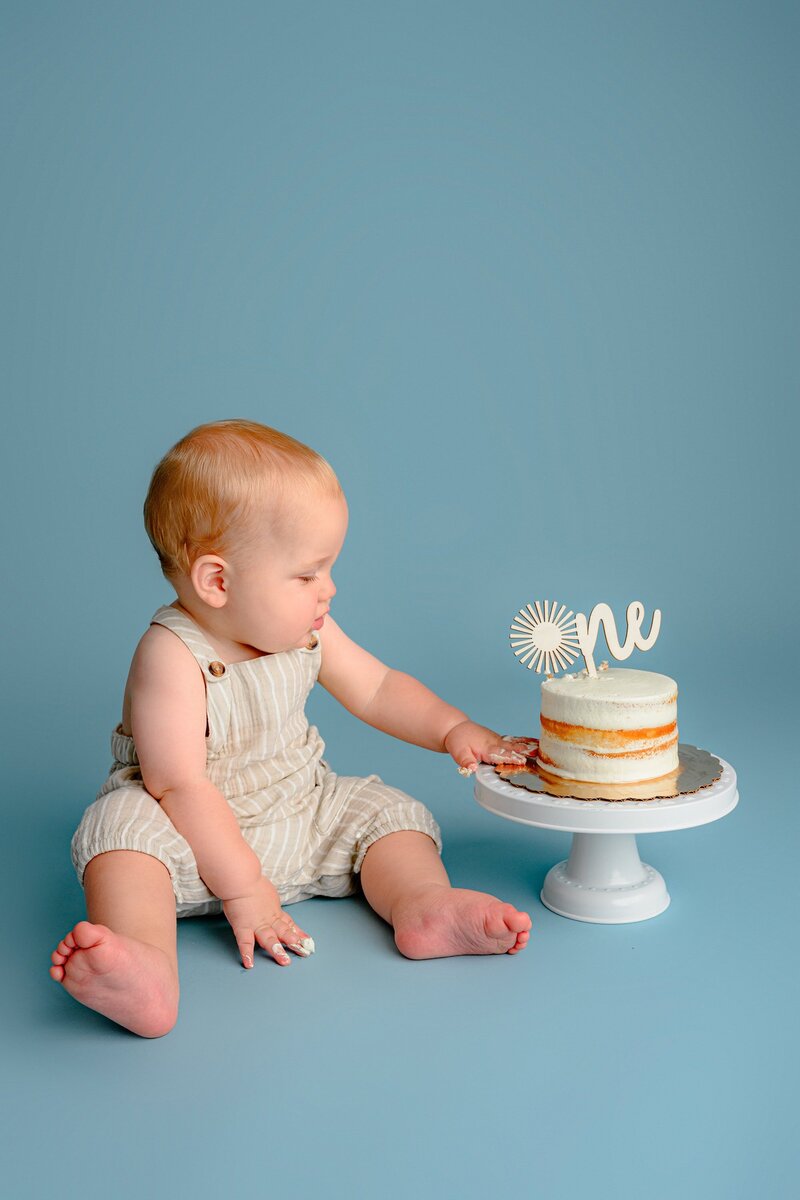 little boy sits in cream overalls on a blue background looking at his smash cake