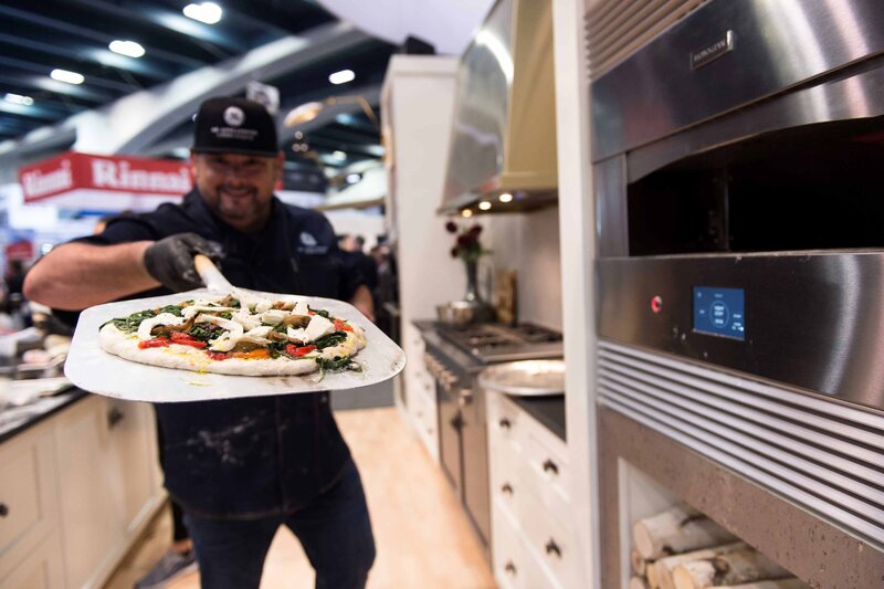 A pizza is displayed at an event 'PCBC trade show to demonstrate the monogram pizza oven