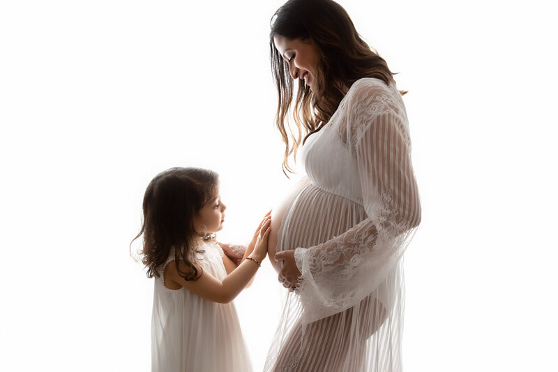 London studio photograph of a pregnant woman in a lace gown with her young daughter lovingly touching her belly.