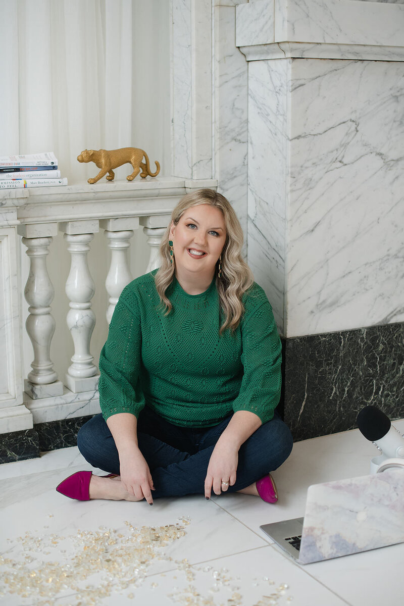 Melissa Lawrence sits on the floor in a green sweater and jeans, surrounded by her laptop and podcast microphone