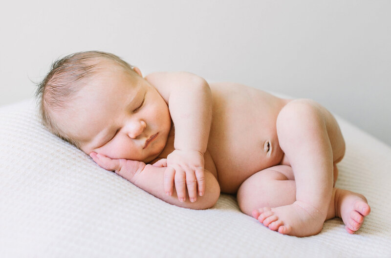 A newborn baby curled up in a cute pose sleeps on a white blanket during a newworn session with Daniele Rose Photography