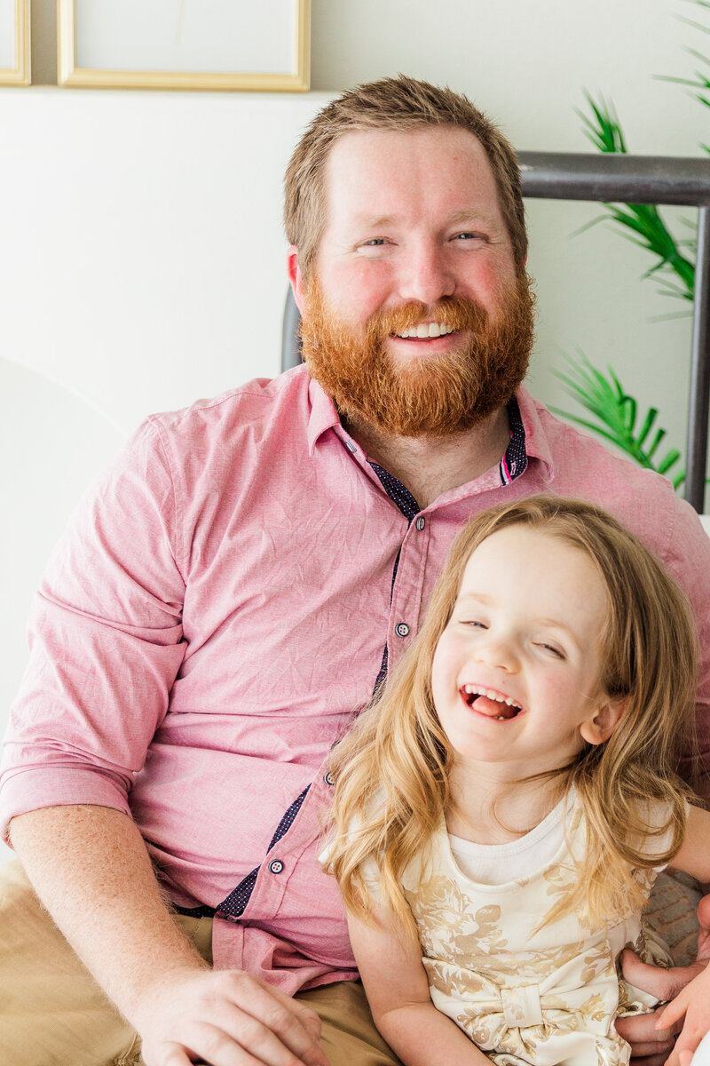 Dad and daughter sit on bed and laugh at camera. Sugarhill Studio Longmont, Colorado