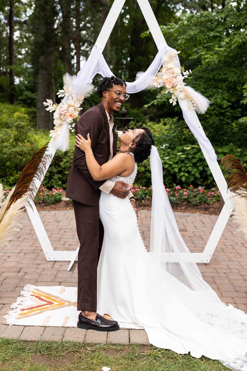 Groom in brown suit with brown leather shoes hugging bride in long trained halter dress with lace detailing hug in front of aframe arbor with plums and dried flowers in brown and tan theme at WRAL azelea garden