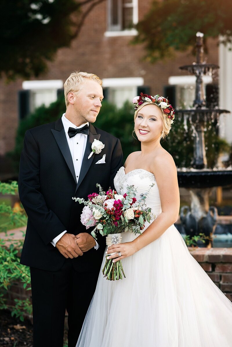Bride and groom standing in front of a brick building. The groom is looking at the bride as she looks at the camera. The groom is wearing a black tuxedo with a white shirt and black bowtie. The bride is wearing a white strapless gown with a sweetheart neckline and a full skirt. The bride is wearing a flower crown that matches her bouquet with blush, deep red, ivory and white flowers and greenery.