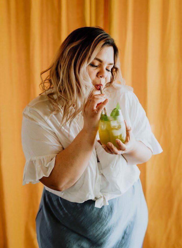 Stephanie Beyer is happily drinking her favourite cocktail out of a straw in front of a yellow velvet backdrop.