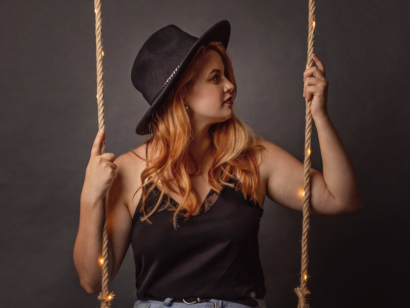 gril in black top and black hat seated on a a lit swing while facing sideways in studio