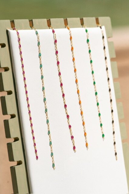 Display of gold-filled enamel chains in multiple colors, including green, pink, orange, and black, arranged on a white jewelry board.