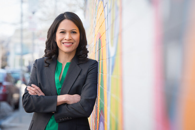 Dr. Jen, a Filipino woman with shoulder-length dark hair, wears a green blouse and dark blazer over top. She is leaning against a colorful mural painted on a brick wall behind her, smiling at the camera.