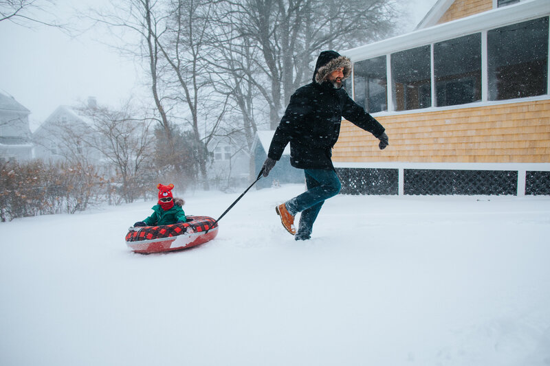 Very bundled up man pulling a very bundled up child on a red sled as snow flies around their faces
