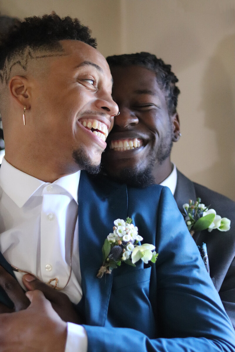 Two Black grooms in blue and gray suits and boutonnieres laugh and embrace