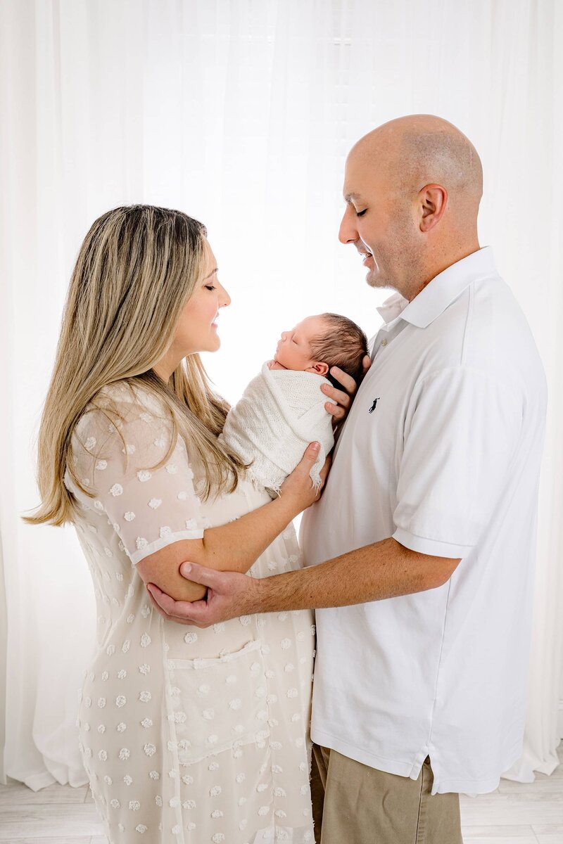 Profile photo of a mother and father holding their newborn son between them with everyone dressed in white in a studio newborn session
