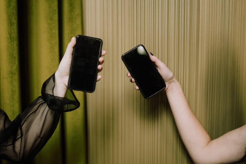 Two hands holding iPhones toast in front of olive green curtains