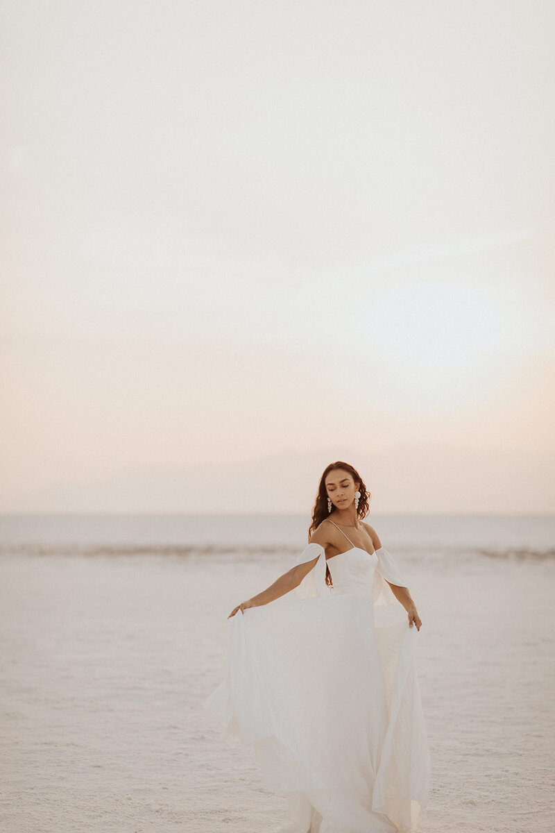 A woman in a flowing white dress stands alone on a sandy expanse.