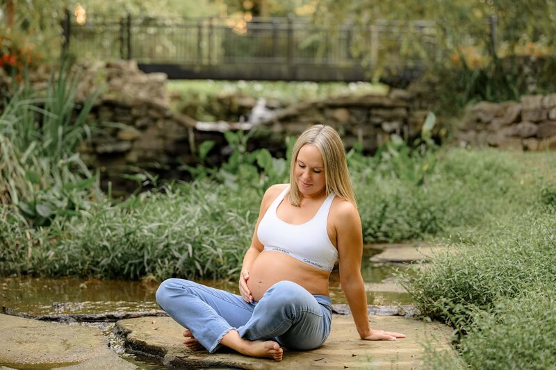 charlotte mother sits in garden setting during maternity session wearing a white sports bra and jeans