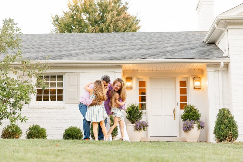 A family of four with two daughters hug in front of their home during a family photo session.