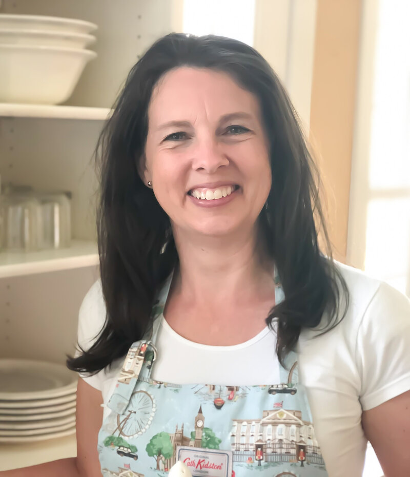 Rachel Sanford in white shirt and Cath Kidston apron standing by white dish cabinet