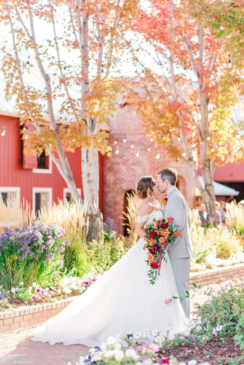 Bride and groom kissing surrounded by colorful flowers and a red barn.