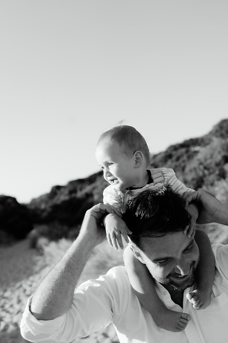 Black and white beach portrait in Geelong showing a smiling father carrying his baby on his shoulders with sand dunes in the background.
