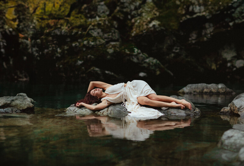 red head woman in a long white gown laying near the water at lynn canyon