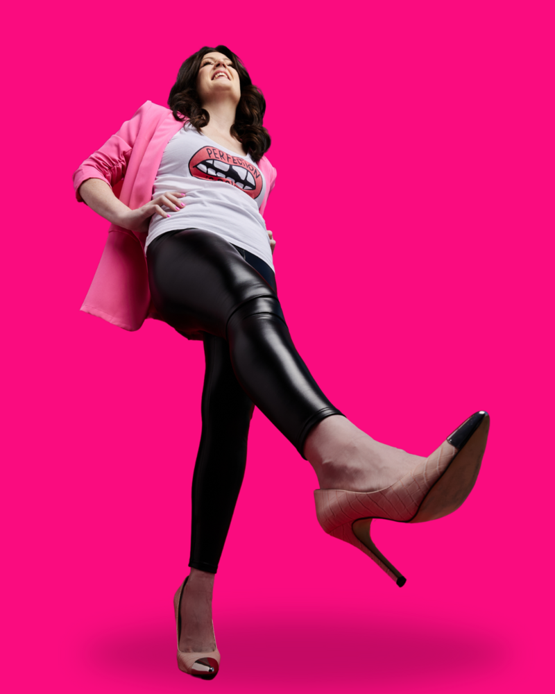 Emily standing wearing light pink heels and kicking her right foot up into the air. She is wearing black pleather leggings and a white shirt with red lips that says perfection is boring and a pink blazer in front of a hot pink background.
