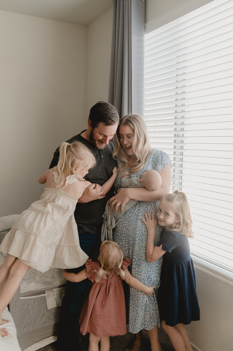 3 sisters snuggle with mom and dad and their newest baby boy next the master window during their newborn portrait session