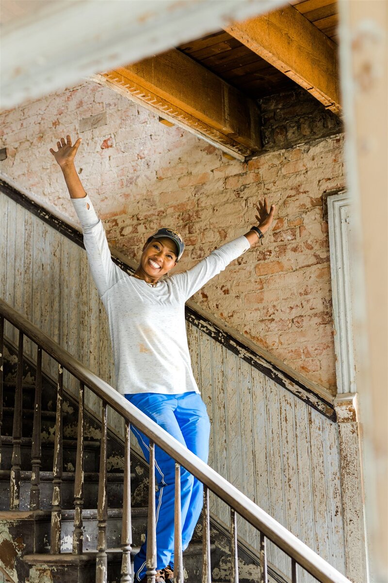 Happy woman standing on the stairs in a hat and blue sweatpants with her arms above her head