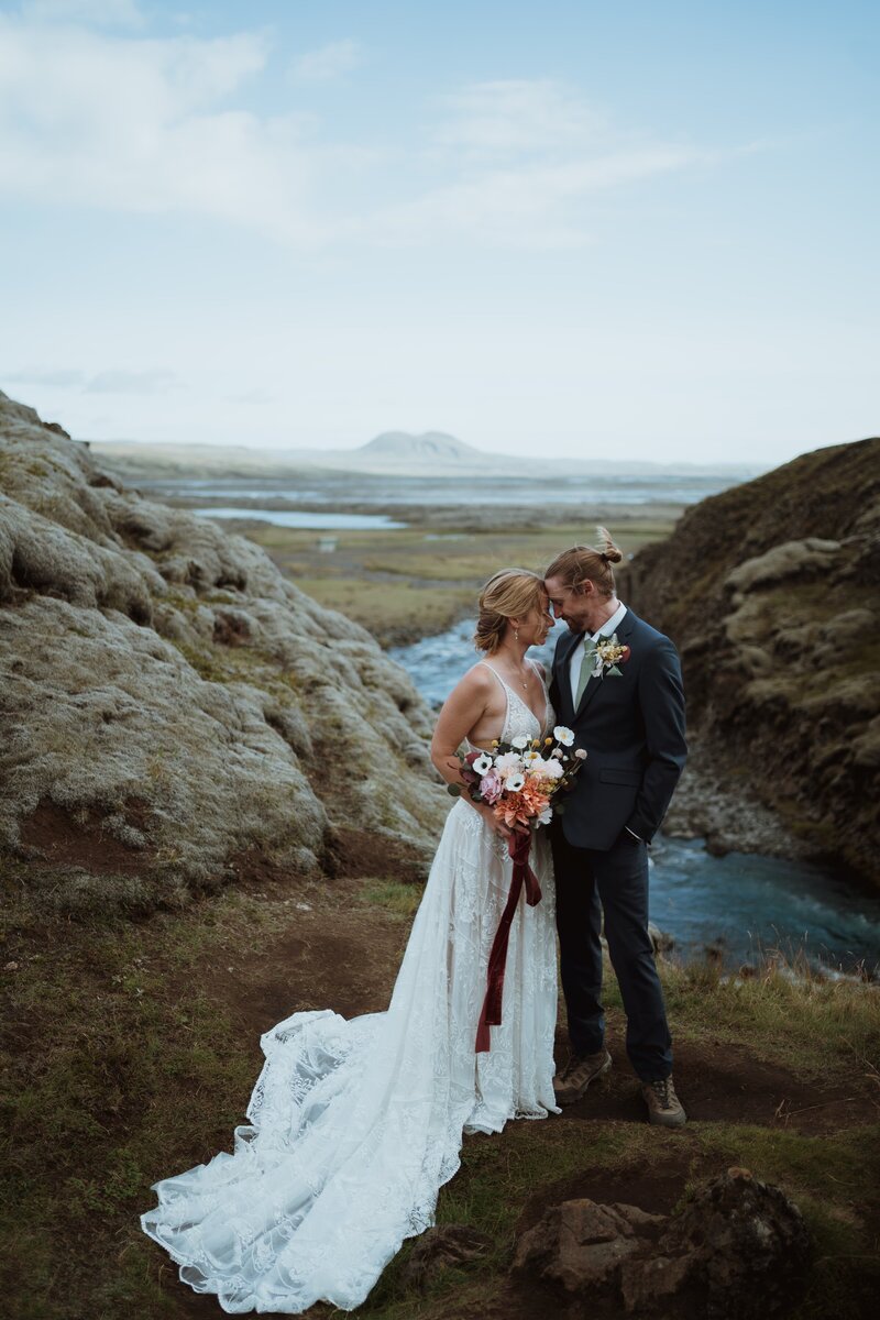 the bride and groom are standing behind a waterfall and have their foreheads pressed up against one another. the bride is holding her flowers up next to her hip and the groom has his hand in his pocket. it is a sunny day.