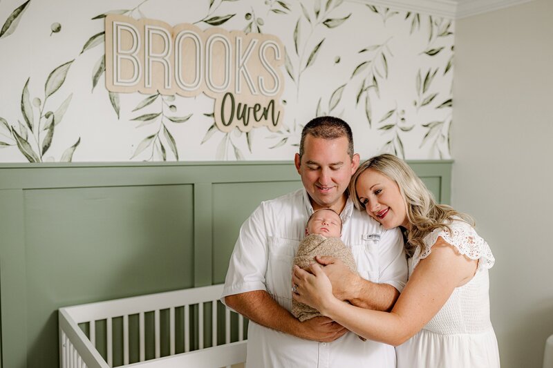 father holds his newborn son wrapped in brown while mom leans against his shoulder in their baby's nursery