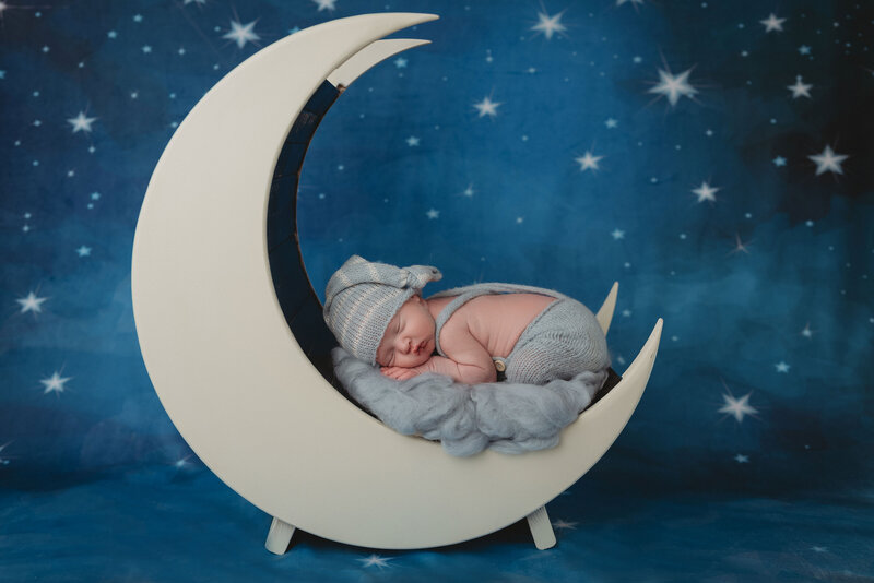 newborn baby boy in blue overalls and  sleepy cap laying on tummy in moon prop with blue starry night backdrop