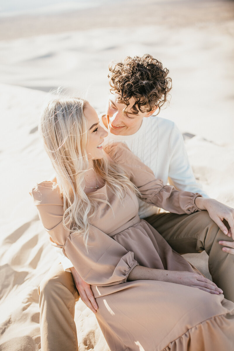 Engaged couple sit together in sand dunes