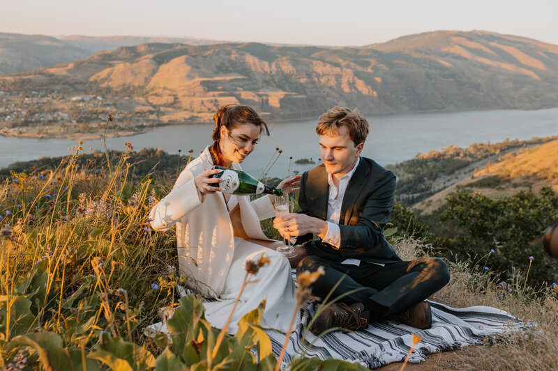 Bride and groom drinking champagne on a picnic blanket overlooking river