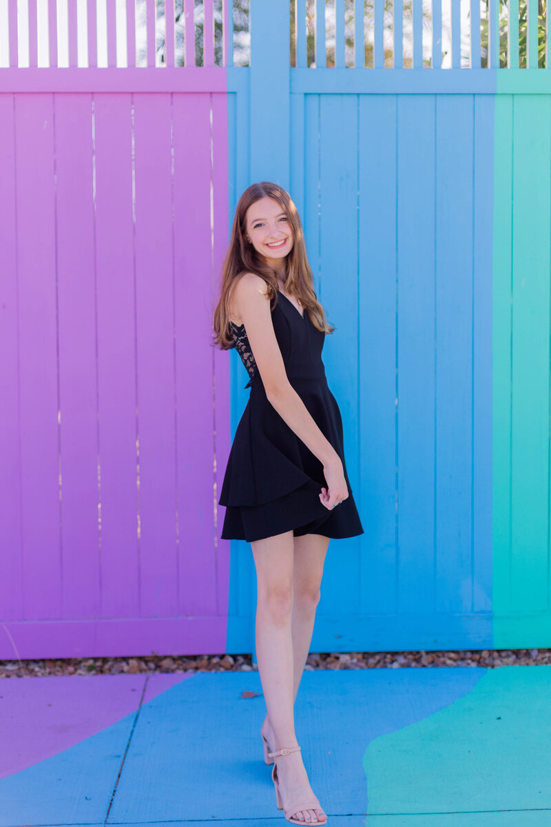 A sixteen year old poses in front of a colorful fence while wearing a black dress on the strand in Galveston