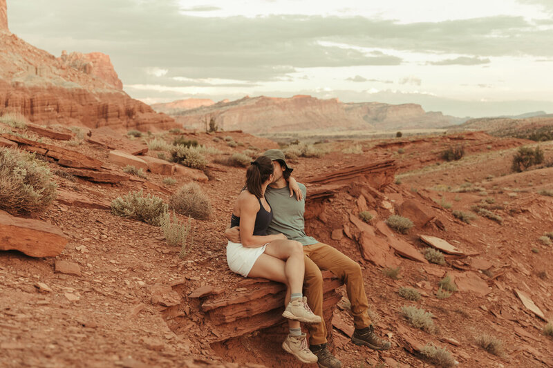 A man and woman in casual hiking clothing, sitting on a red rock ledge in Torrey, Utah, kissing so you cannot see either of their faces. The beautiful red rocks of Capitol Reef are behind them.