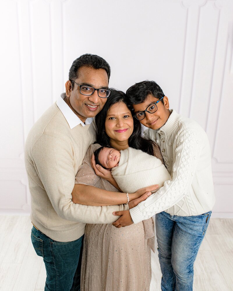 Mother cradle's newborn son with both her partner and her oldest son beside her all dressed in neutral tones with the baby wrapped and cream in a Charlotte Area Studio