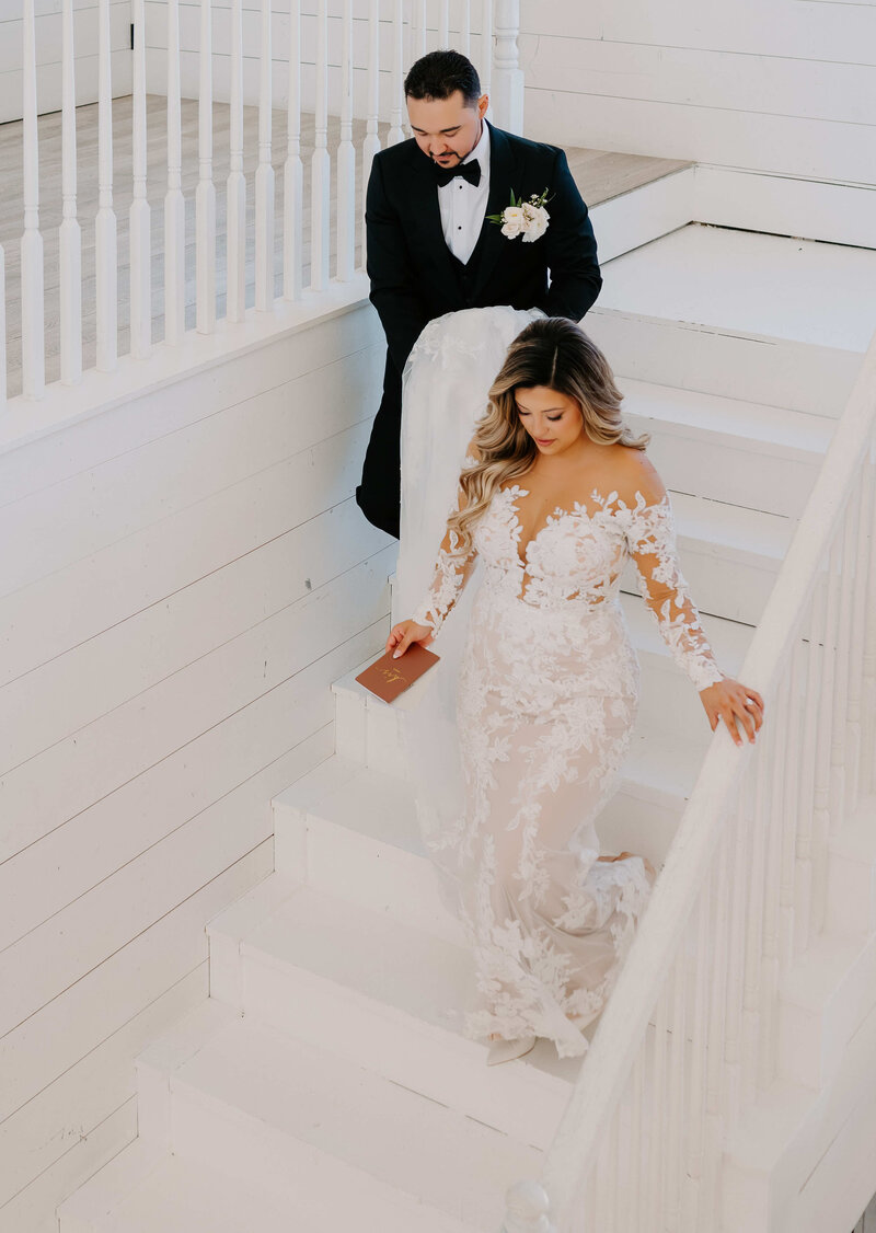 A bride in a white lace wedding dress and a groom in a black tuxedo and bow tie are walking down a set of white stairs.