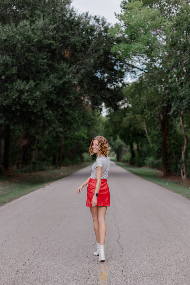 Hopeful Katy Texas senior turns to look at camera on an empty street while wearing a red leather skirt white top and boots