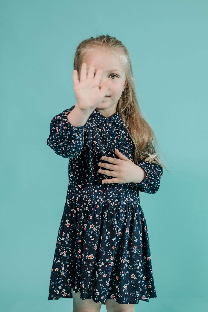 A young girl in a floral dress raises her hand towards the camera against a teal background, partially covering her face.