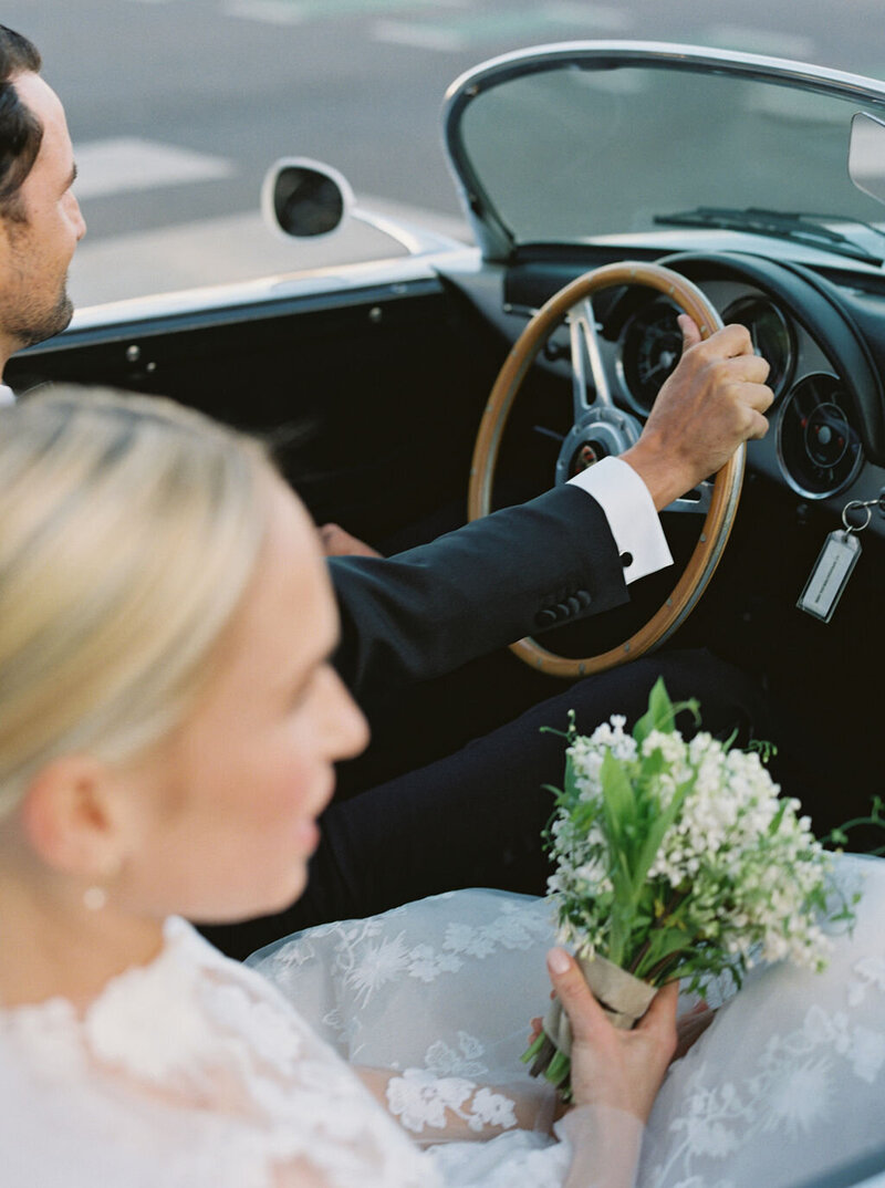 Image taken over the bride's shoulder. Bride and groom driving away in a convertible.