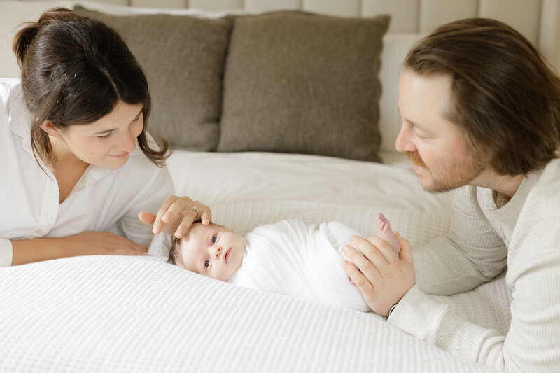 Mom and Dad smiling at their newborn baby on bed in their newborn session with Caroline Eidson