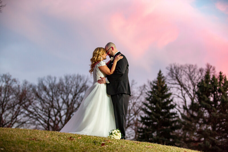 A bride and groom posing on a hill with dramatic sunset on the background.