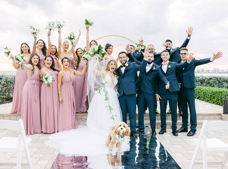 a bride and a groom surrounded by bridesmaids dressed in pink and groomsmen dressed in dark blue suits with their hands up and a small fluffly dog in front of them