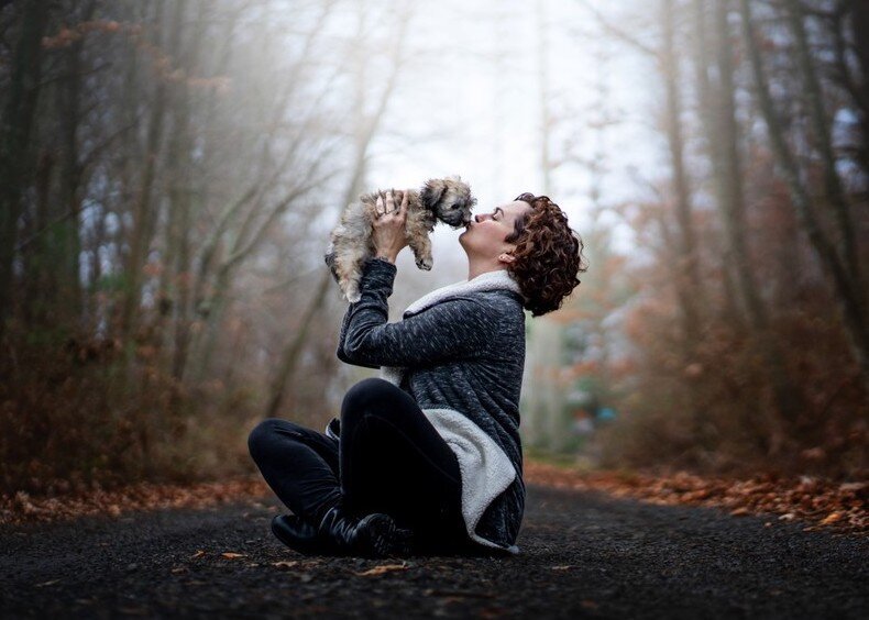 A woman sitting on the ground holding up a small dog and kissing it.