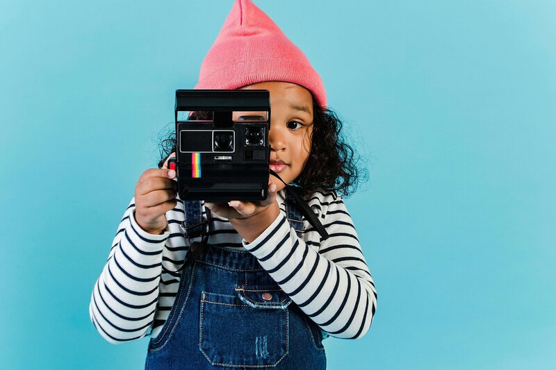 A child wearing a pink hat, striped shirt, and denim overalls is holding a vintage Polaroid camera, looking through the viewfinder against a light blue background.