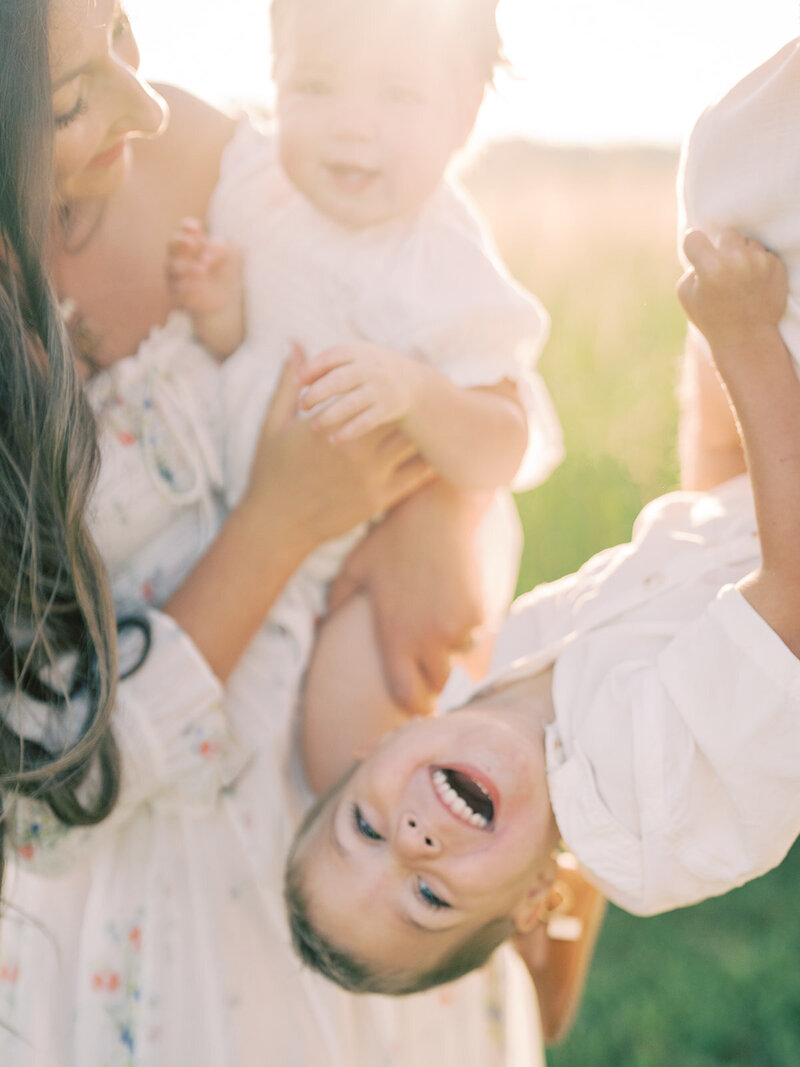 Little boy is held upside down by father as mother stands next to them holding toddler daughter while laughing, photographed by Northern VA Family Photographer Marie Elizabeth Photography