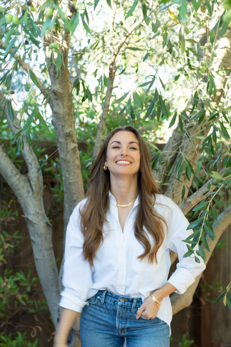 A photo of Tara Accardo, with long brown hair who is smiling in a white button up shirt, from Losses Become Gains