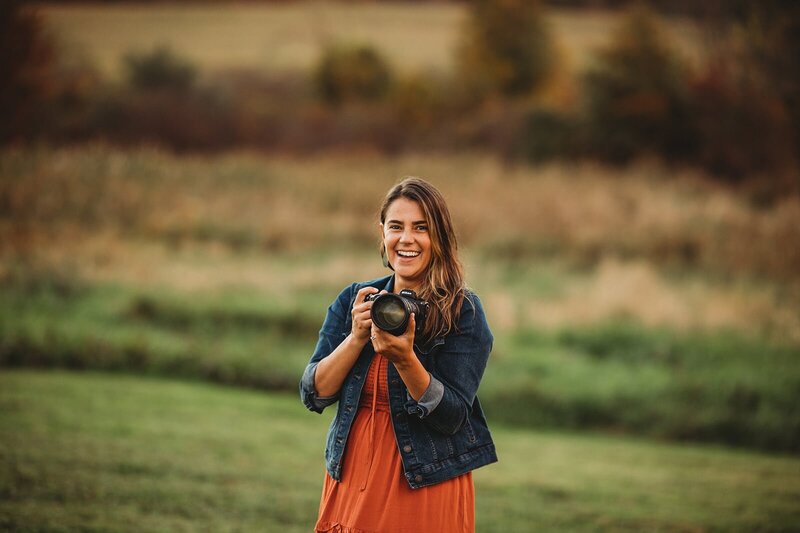 A happy woman in a field during golden hour, wearing a denim jacket over an orange dress, holding a camera with a joyful expression.
