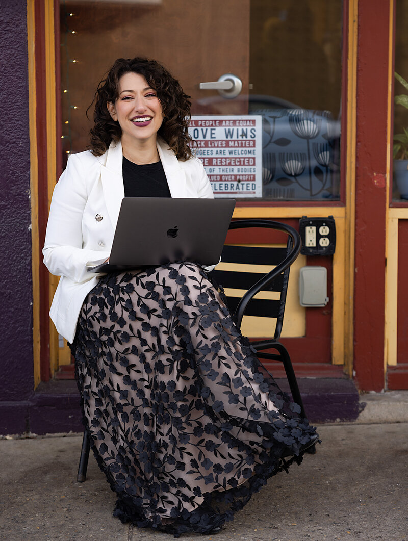 Woman in a floral skirt and white blazer sitting in a chair with open laptop smiling at the camera