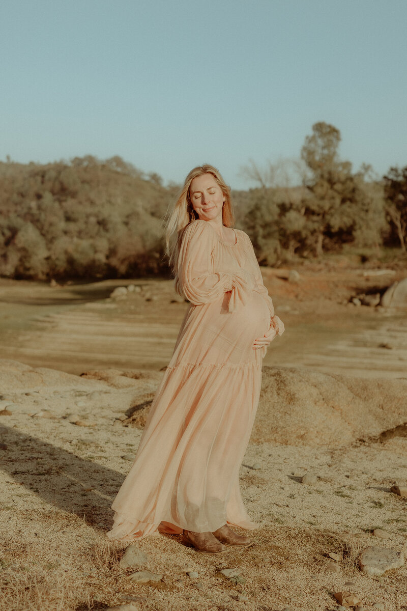 A pregnant woman in a long dress standing on a rock by a river.